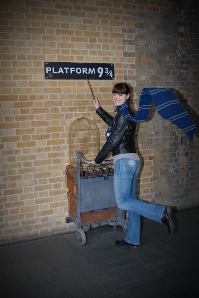A woman poses with a luggage cart at the 'Platform 9¾' sign, holding a wand and wearing a blue scarf.