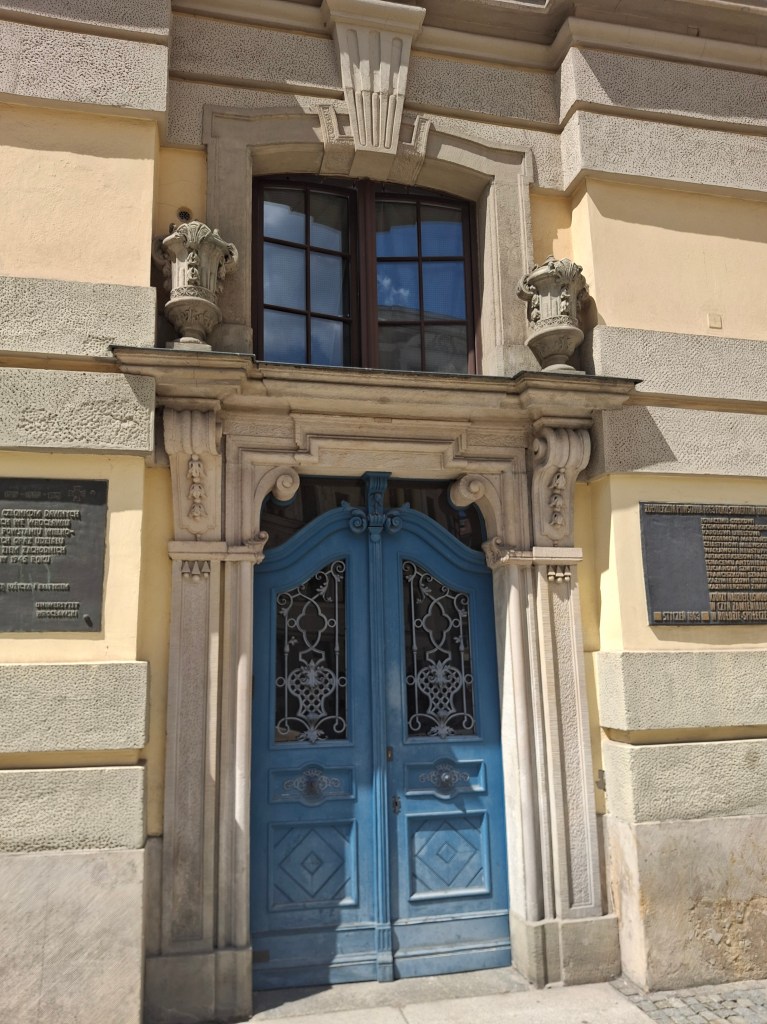 An ornate blue door with decorative metalwork, framed by stone columns and a detailed archway, set against a yellow wall and accompanied by decorative urns and plaques.