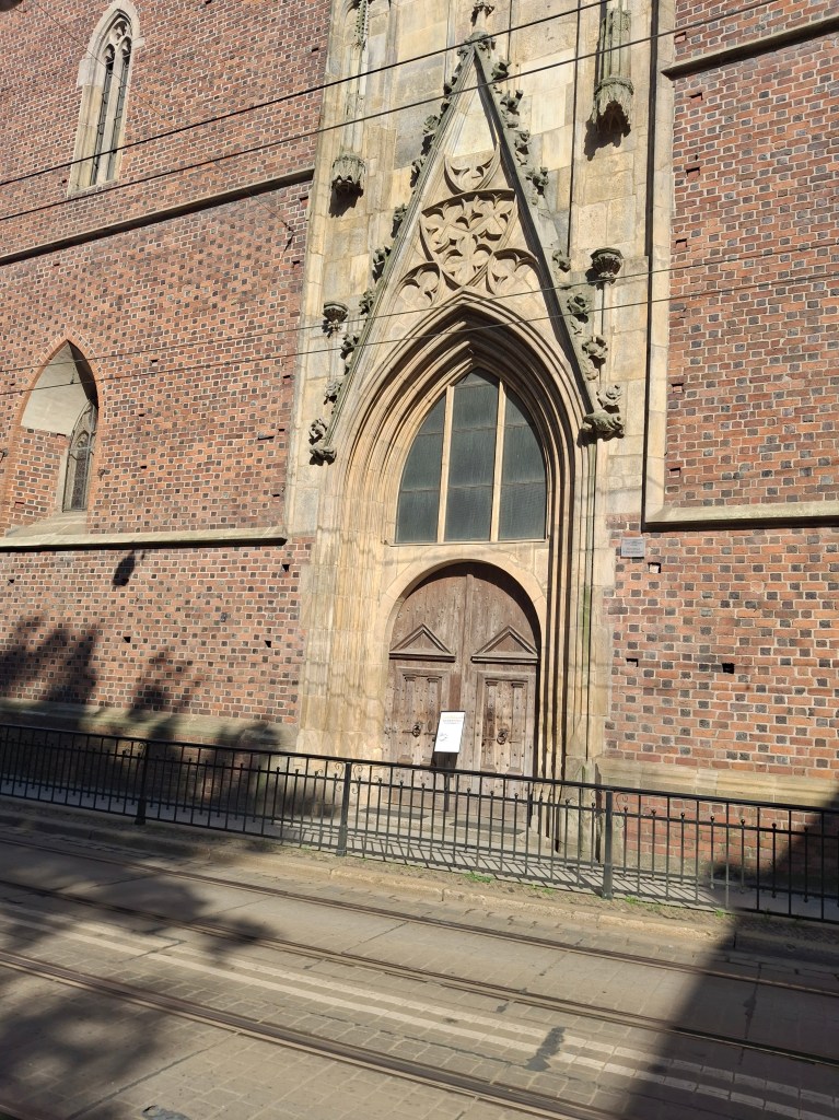 Exterior view of a historic brick building featuring a large archway with decorative stonework and wooden doors, flanked by small windows and surrounded by a metal railing.