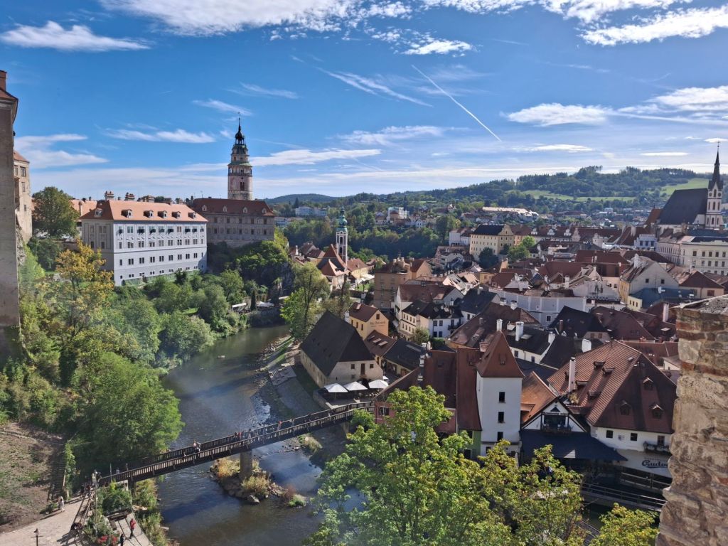 Panoramic view of a charming town with medieval architecture, featuring a river, a bridge, and hills in the background under a clear blue sky.